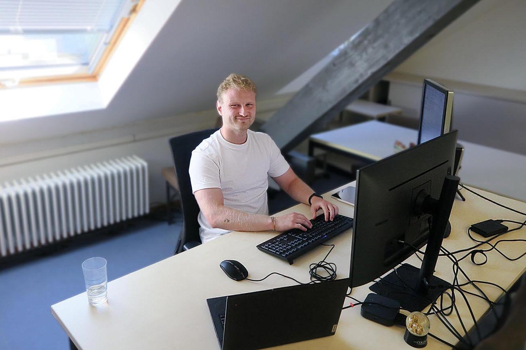 Eine Person sitzt am Schreibtisch und lächelt in die Kamera.